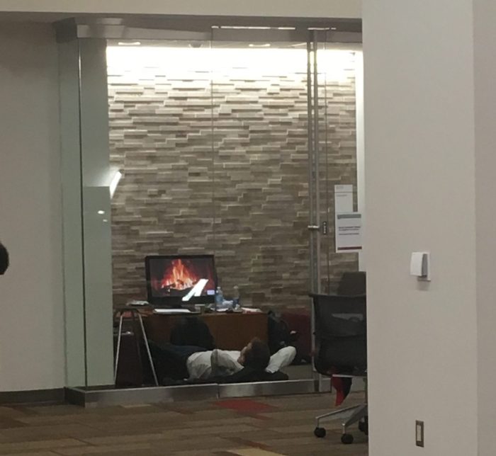 student lying on the ground watching a screen with fireplace on it in group study room